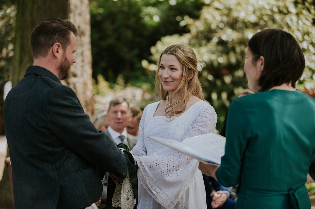 A white opposite sex couple face each other during their wedding ceremony. Their hands are bound with two Handfasting cords - tartan and lace. They are smiling. 