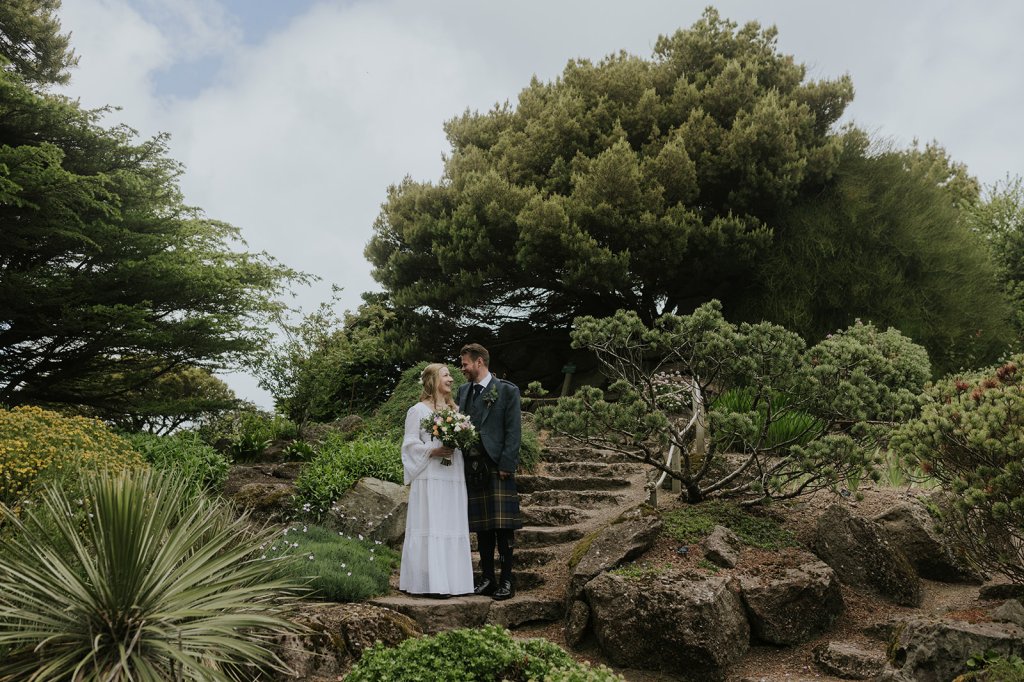 A white opposite sex couple stand on a stone staircase in a garden. They are flanked by trees and shrubs and wearing wedding clothes. She is wearing a long white wedding dress and holding a spring bouquet of flowers. He is wearing a kit and tweed jacket. They are smiling at each other. 