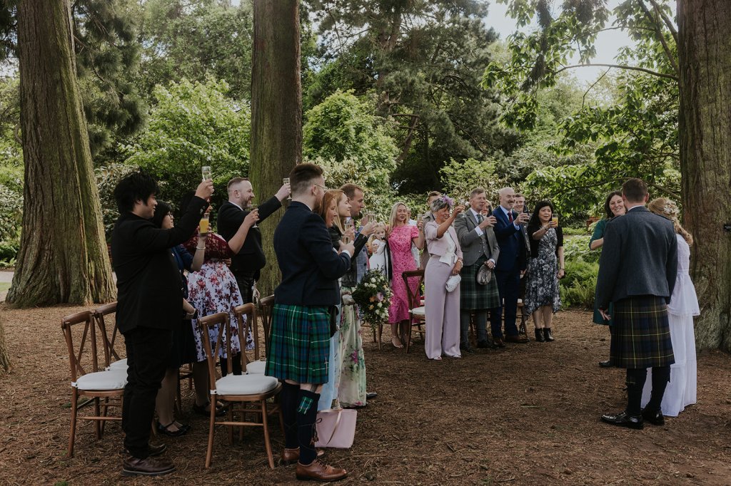 A wedding party stand circled by ancient rewoods in a botanic garden. They are raising a toast to two newlyweds. Everyone is smiling. 