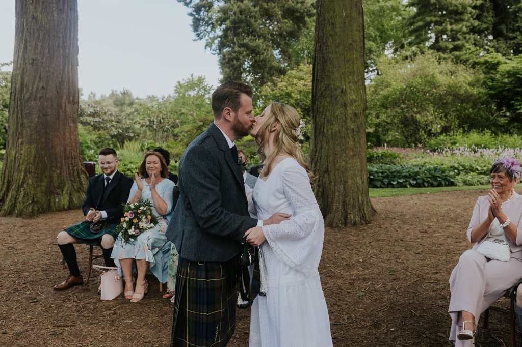 A white opposite sex couple kiss during their wedding ceremony. He is wearing a tartan kilt and blue tweed jacket. He has short dark hair and a small beard. She has blonde hair and is wearing a white dress with bell sleeves and long skirt. Their loved ones look on smiling and clapping. 