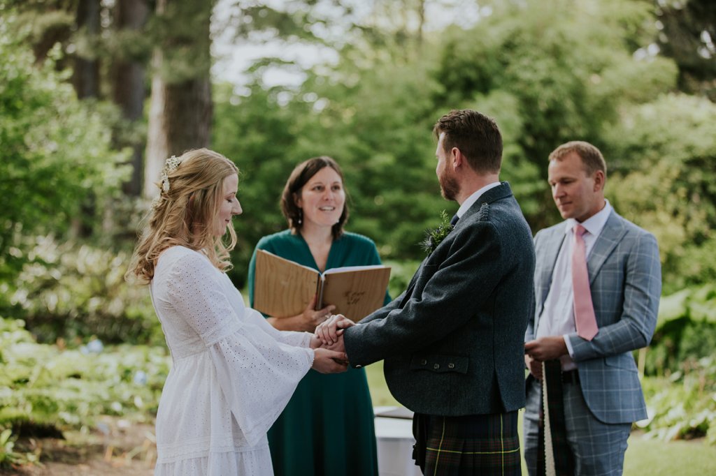 A white opposite sex couple hold hands facing each other during their wedding ceremony. She's wearing a white dress with long bell sleeves and floor length skirt. He's wearing a tartan kilt and blue jacket. She has long blonde hair and he has short brown hair and a beard. The celebrant stands between them holding a wooden folder with the words 'love is all' written on it. The celebrant is smiling. To their right a man with a grey checked suit, pink tie and white shirt with cropped blonde hair holds their Handfasting ribbons - a piece of tartan and a piece of white lace.