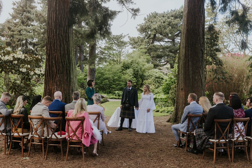 A white opposite sex couple stand facing an intimate circle of their loved ones beneath two towering redwoods. They are dressed in wedding clothes. He's in a kilt and jacket and she's in a white dress. Their family sit on rows of wooden chairs. The celebrant stands to the left of the couple in a green dress sharing their story. 